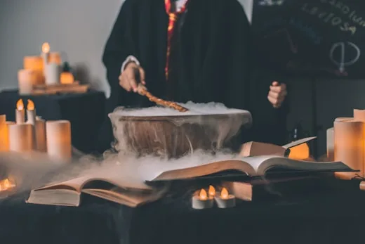 A cauldron bubbles with a potion as someone casts spells from a spellbook, showcasing creative arts.